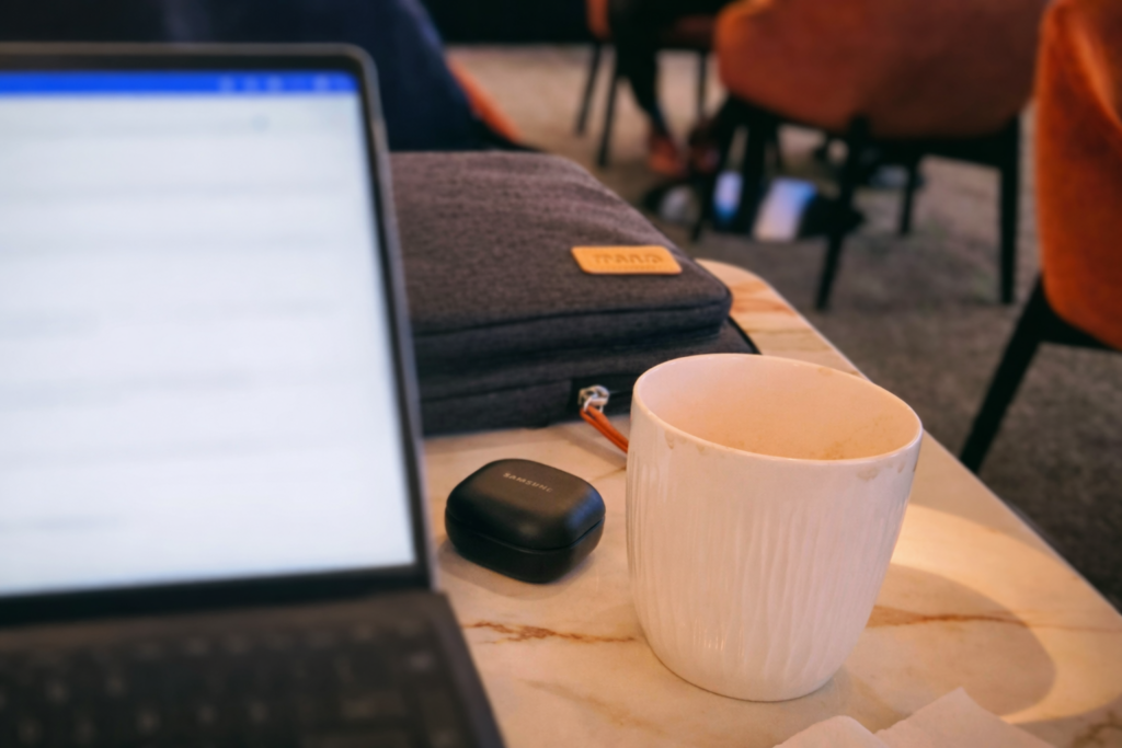 A laptop sits on a marble café table next to a white coffee mug, a fabric tech pouch, and wireless earbuds, with chairs blurred in the background.