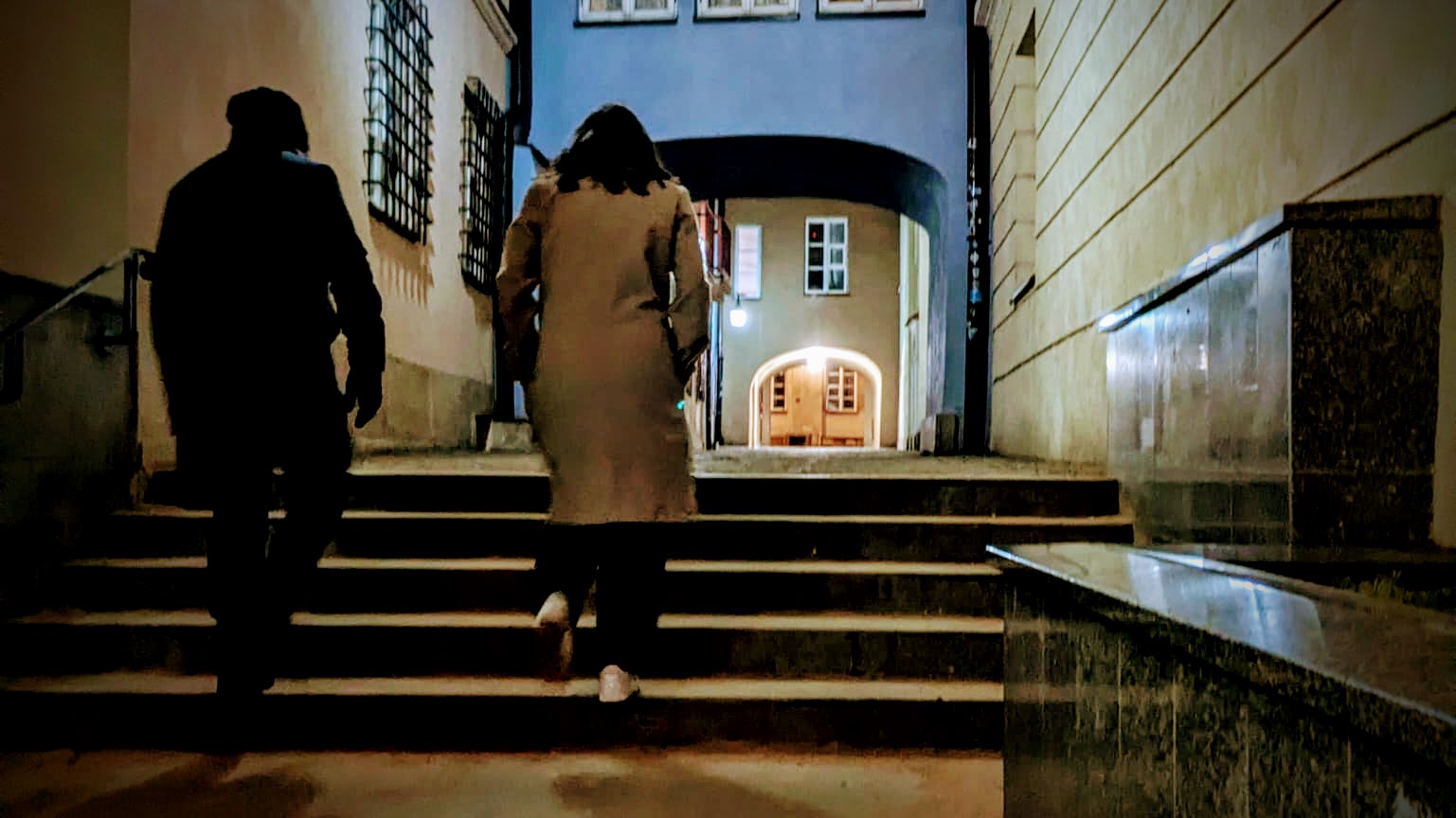 Man and woman walking up stone steps in a narrow historic Polish alley at night toward a softly lit archway.